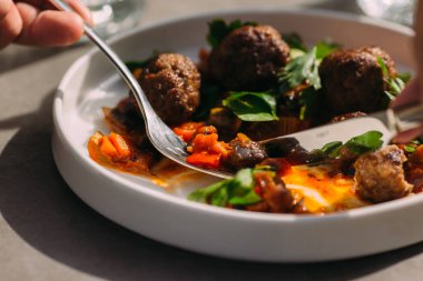 Çatal ve bıçak kullanan, soslu köfte ve sebzelerden hoşlanan, doğal ışığı olan bir restoranda yemek yiyen biri.