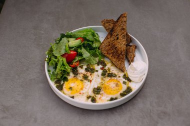 Plate of fried eggs topped with pesto, fresh green salad, toasted bread, and cream on a gray background