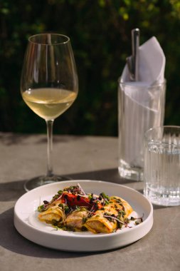 White plate of pesto pasta rolls garnished with fresh sprouts, served with a glass of chilled white wine on a sunlit terrace