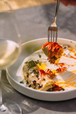 Hand holding a fork with a piece of savory pancake with sauce, next to a wine glass on a outdoor table