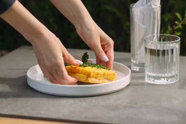 Eller iştah açıcı bir sandviçi beyaz bir tabağa taze yeşillikler ve açık bir masada bir bardak su ile yerleştiriyor.