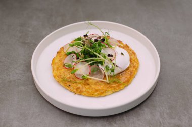 Savory vegetable pancake on a white plate, topped with a poached egg, sliced radish, and fresh microgreens, ready to eat