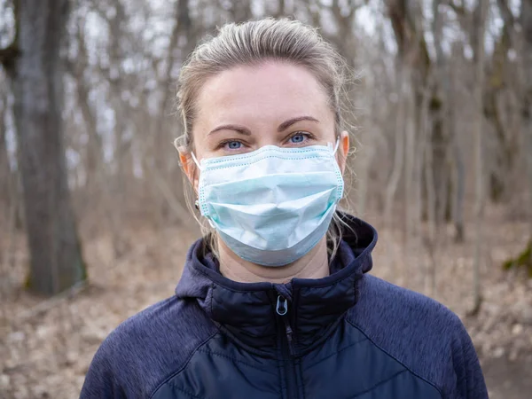 Mavi ya da yeşil renkli tıbbi maske takan sarışın güzel bir kadının yüzü. Parkta bir kız yürüyor. Koronavirüs salgını sırasında sağlığın korunması. Sağlıklı yaşam tarzı kavramı.