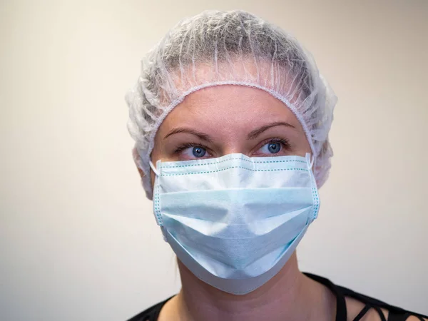 Tıbbi mavi maske takan genç bir kız. Başında koruyucu bir şapka ya da şapka var. Model, açık renkli duvar arka planında izole edilmiş. Karantina ve özgürlük kısıtlaması kavramı. Yakın plan.