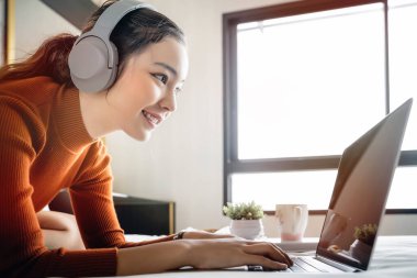 Kulaklıkla müzik dinleyen ve evde dinlenirken dizüstü bilgisayar kullanan neşeli genç bir kadının portresi..