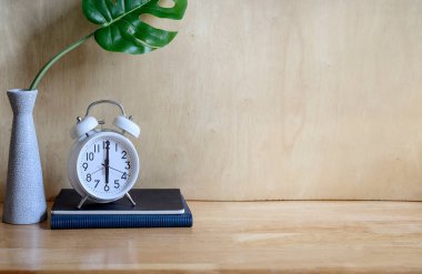 Tahta masadaki ev bitkisi ve kitap üzerindeki beyaz alarm saatini taklit et, ürün gösterimi veya montaj için alanı kopyala.