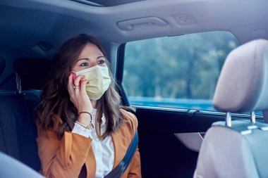 Yüzü maskeli beyaz bir kadın cep telefonunu arabada takıyor ve iş kadını cep telefonuyla konuşuyor.