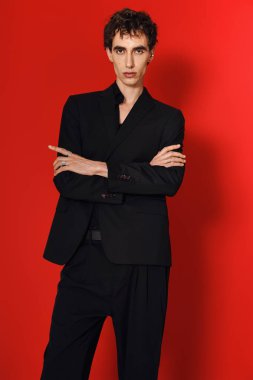 Zarif siyah giyen yakışıklı bir adam kollarını kavuşturup kırmızı bir arka plana yaslanıyor..