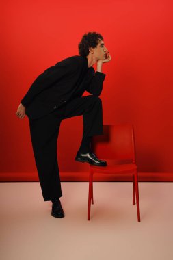 Zarif siyah elbiseli yakışıklı adam bacağı kırmızı sandalyede düşünceli bir şekilde poz veriyor..