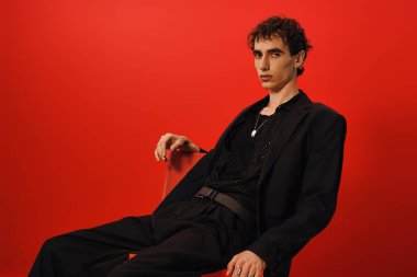 Şık bir adam otururken zarafet sergiler, parlak kırmızı bir arka planda keskin bir şekilde giyinir..