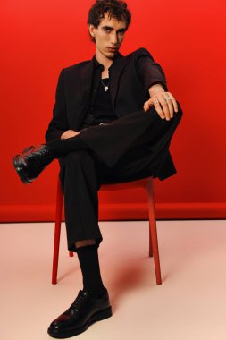 Parlak siyah elbiseli yakışıklı adam, koyu kırmızı bir arkaplana karşı güvenle poz veriyor..