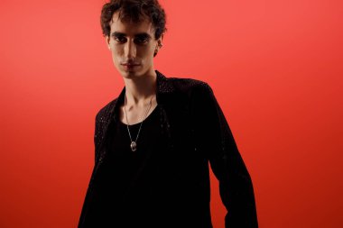 Yakışıklı adam şık kıyafetler sergiliyor, koyu kırmızı arka planda güvenle duruyor..