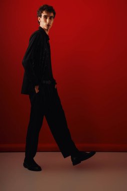 Parlak siyah elbiseli yakışıklı adam çarpıcı bir kırmızı arkaplan önünde güven içinde poz veriyor..