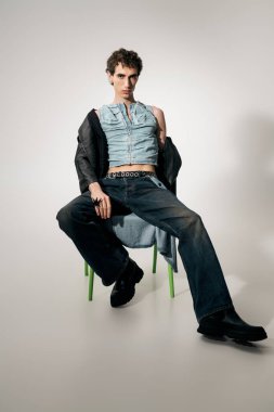 Tarz sahibi bir adam yeşil bir sandalyede kendine güvenerek poz veriyor, eşsiz bir moda karışımı sergiliyor..