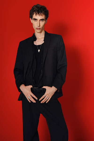 Стильный человек уверенно стоит, источая очарование в гладкой одежде против дерзкой красной обстановке.