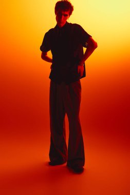 Handsome young man stands confidently in fashionable clothing, illuminated by warm orange light.