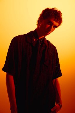 Handsome young man showcases modern fashion while standing in front of a warm orange backdrop.