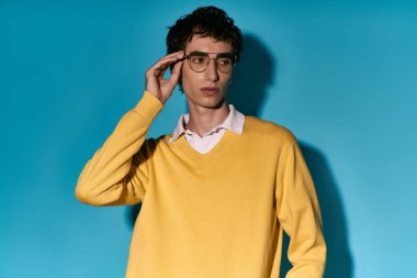 A dapper young man in a yellow sweater poses confidently against a bright blue backdrop.