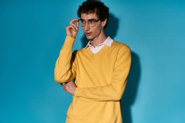 A young man confidently poses in a trendy yellow sweater with glasses on a vibrant teal background.