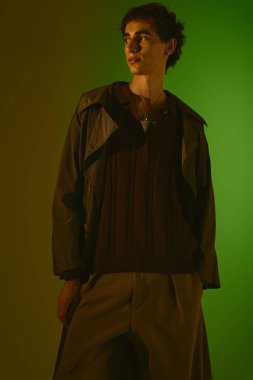 A handsome young man stands confidently in fashionable clothing, illuminated by green light.