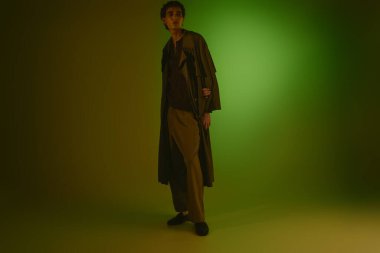 Stylish young man poses confidently in trendy attire amidst a striking green background.