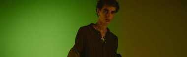 A handsome young man presents unique attire against a striking green backdrop, exuding confidence.