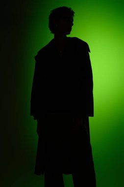 A confident young man stands silently, dressed in fashionable attire, illuminated by a green light.