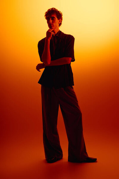 Handsome young man in trendy attire poses thoughtfully with a warm gradient background.
