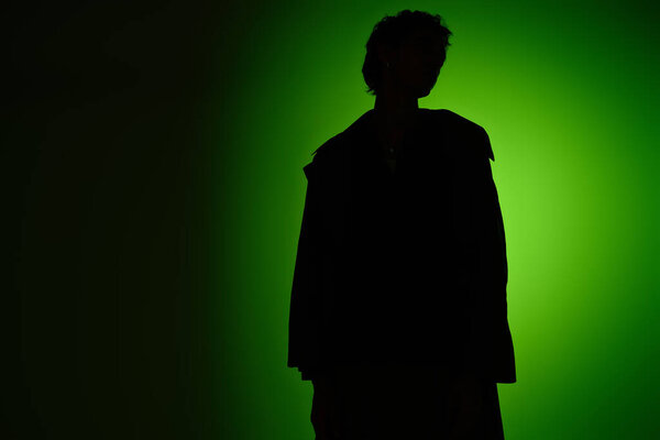 A handsome young man poses in silhouette against a vibrant green background, showcasing fashion.