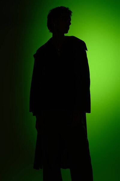 A confident young man stands silently, dressed in fashionable attire, illuminated by a green light.