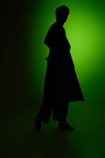 A handsome young man walks gracefully in stylish attire, silhouetted by a bright green light.