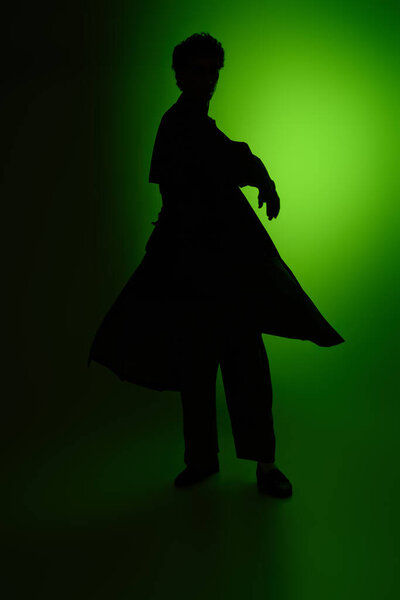 A young man dressed in stylish clothing strikes a bold pose, silhouetted by a green light.