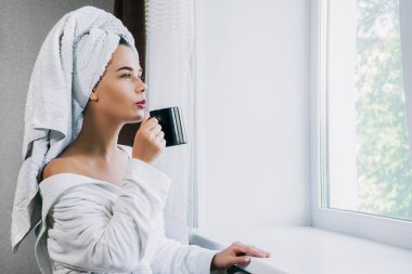 Beyaz havlu ve bornozla genç ve güzel bir kadın evdeki pencerenin yanında kahve içiyor.