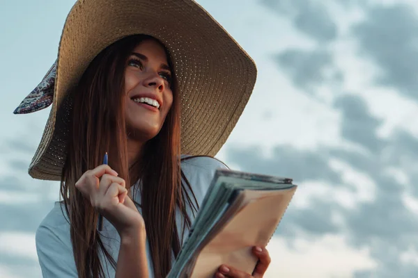 Young woman in straw hat writing in a notebook at nature. Inspiration concept