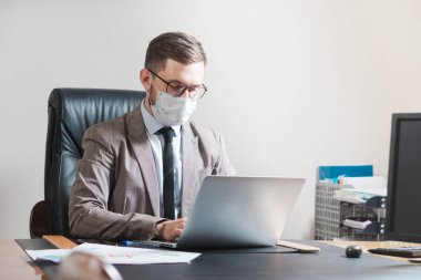 Gözlüklü ve maskeli genç işadamı kişisel ofisinde dizüstü bilgisayarla çalışıyor. Karantina sırasında iş.