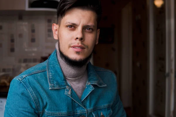 Young nice bearded man in jean jacket and roll-neck sweater at home.