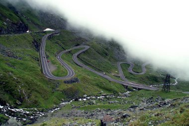 Romanya 'nın Balea nehri vadisindeki Transfagarasan yolu. Yağmurlu bir yaz gününde Balea nehri boyunca bulutların altında uzanan dolambaçlı bir yol..