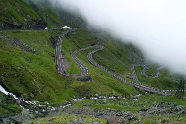 Romanya 'nın Balea nehri vadisindeki Transfagarasan yolu. Yağmurlu bir yaz gününde Balea nehri boyunca bulutların altında uzanan dolambaçlı bir yol..