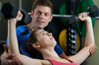 Woman In Gym Lifting Weights Encouraged By Personal Trainer clipart