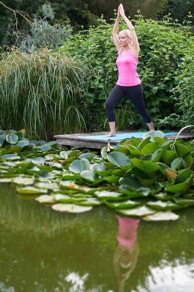 Mulher madura na posição de Yoga no molhe de madeira pelo lago — Fotografia de Stock