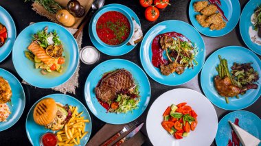 Masadaki farklı yemeklerin üst görüntüsü. Çorba, kızarmış hamburger, ızgara sebze, soslu kızarmış tavuk budu ve salata.