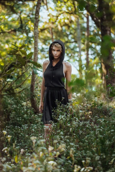 Brütende verträumte junge Blondine, schwarzes Kleid mit Kapuze am Baum stehend — Stockfoto