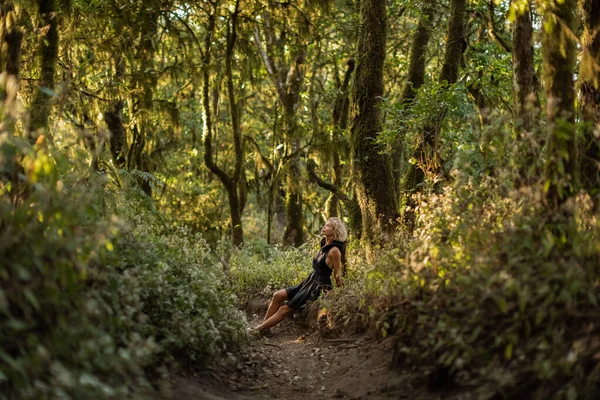 Jong meisje blond, zwart jurk met een kap lopen in het bos zitten in een gras — Stockfoto