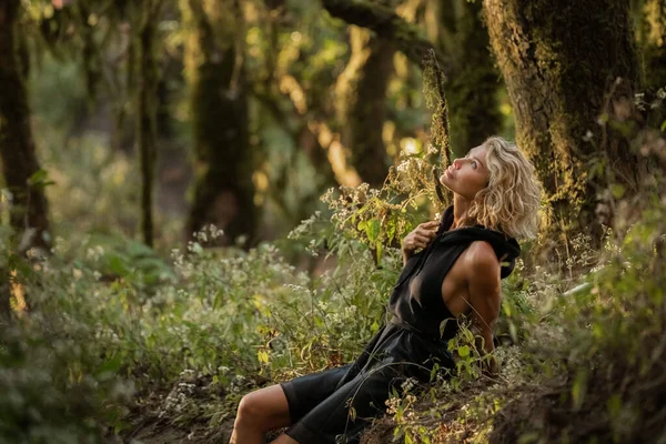 Blond, svart kjole med hette går i skogen og sitter i gresset – stockfoto