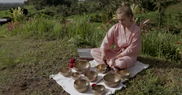 Blonde woman in a pink kimono playing on meditative Himalayan singing bowl. — Stock Video