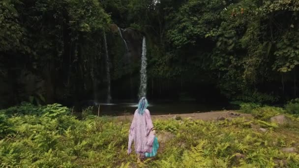 Жінка в рожевій сукні йде до високого водоспаду в тропічних джунглях Індонезії. — стокове відео