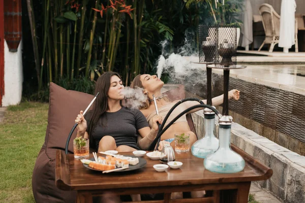 Group of young girls smoking a hookah and exhaling white smoke. Vacation.