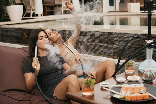 Group of young girls smoking a hookah and exhaling white smoke. Vacation.