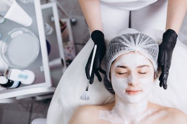 Yüzünde spa ameliyatı olan genç bir kadın. Spa masasına uzanmış bir kadın ve kozmetik maske takan bir kozmetik uzmanı. Metnin için boşluk kopyala