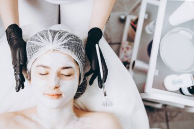 Yüzünde spa ameliyatı olan genç bir kadın. Spa masasına uzanmış bir kadın ve kozmetik maske takan bir kozmetik uzmanı. Metnin için boşluk kopyala